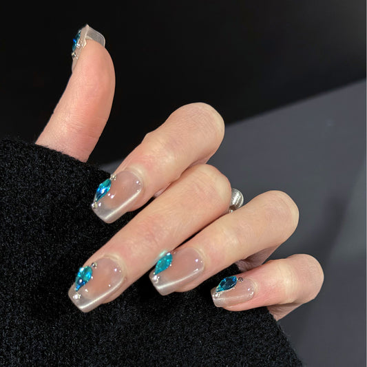 Hand model wearing Aqua Drop press-on nails with sheer nude cat-eye finish and aqua gemstone trim, showcasing the shimmering effect.